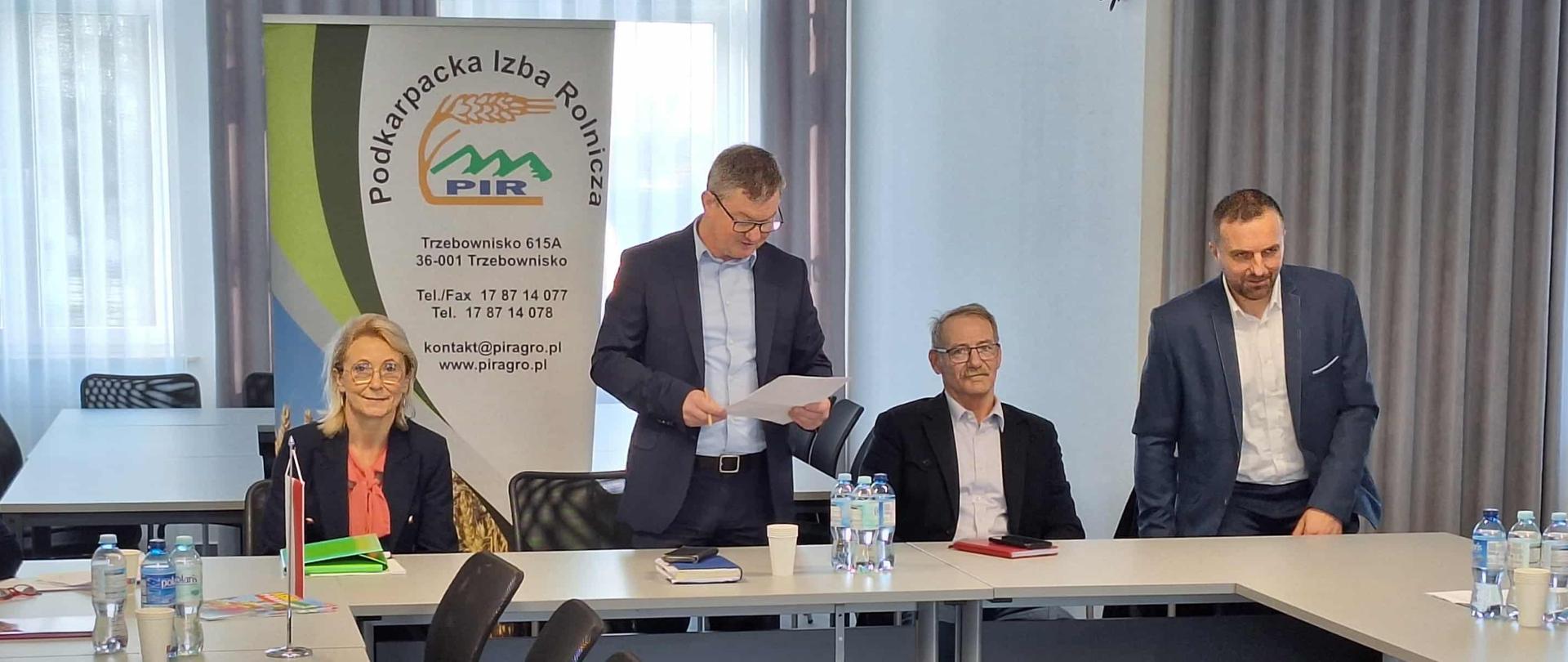 Cztery osoby siedzące przy jasnym stole. Jedna kobieta i trzech mężczyzn. Jeden z nich stoi i trzyma w dłoni papier. W tle stoi baner z logiem instytucji. Na stole stoi woda w butelkach oraz kubki.