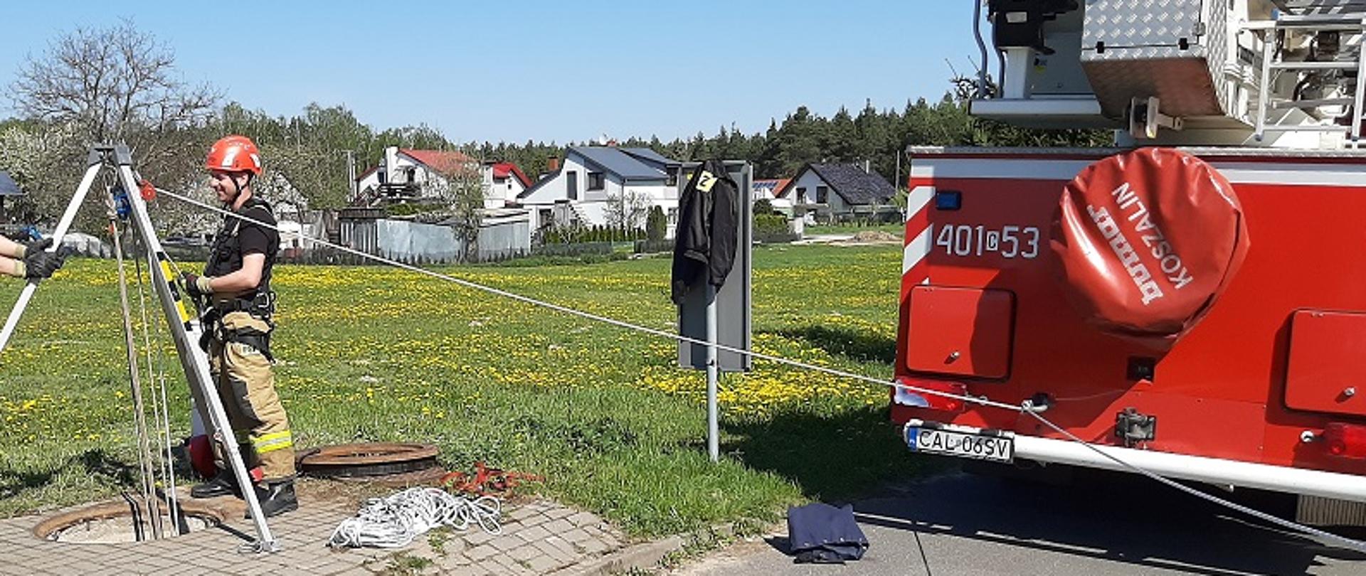 Na zdjęciu widać dwóch strażaków. Jeden ubrany jest w czarne buty, spodnie, koszulkę na krótki rękaw oraz czerwony hełm, drugi posiada czarne buty i koszulkę na krótki rękaw, spodnie koloru piaskowego i czerwony kask alpinistyczny. Funkcjonariusze stoją przy srebrnym trójnogu ratowniczym rozstawionym na szarej kostce brukowej nad otworem studni. Do trójnogu podłączone są niebieski i czerwony bloczek ratowniczy oraz białe liny. Jedna z nich poprowadzona jest do czerwonego samochodu strażackiego stojącego po prawej stronie od ratowników. Widoczny jest jedynie tył samochodu ciężarowego a na nim po środku pokrowiec koła zapasowego z napisem BUMAR KOSZALIN. Po lewej widnieje napis 401C53. Litery są koloru białego. Nad napisami widnieją srebrnego koluru elementy podnośnika hydraulicznego zamontowane na pojeździe. Przed strażakami widoczny jest fragment szarego asfaltu, po lewej zaś stronie część skrzynki elektrycznej. Tło zdjęcia stanowi zielona trawa z żółtymi mniszkami lekarskimi, drzewa liściaste, domy o jasnych elewacjach i ciemnych dachach oraz bezchmurne niebo.