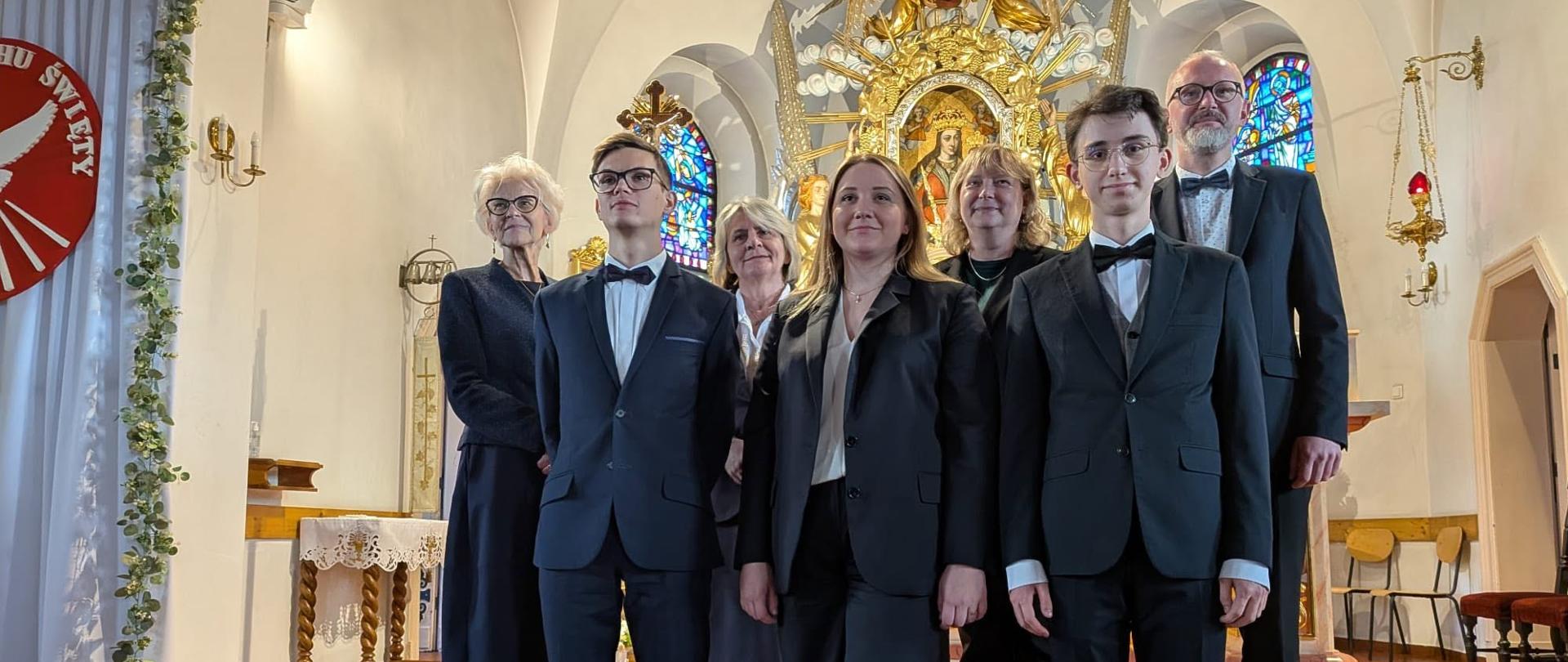 Zdjęcie grupowe muzyków w kaplicy. Sześcioro osób ubranych w eleganckie czarne stroje stoi w rzędzie we wnętrzu kaplicy o jasnych ścianach i sklepieniu. Od lewej: chłopiec w garniturze z muszką, mężczyzna w garniturze z muszką, kobieta w czarnym stroju, kobieta w czarnym stroju, młody mężczyzna w garniturze z muszką oraz starszy mężczyzna w garniturze. W tle widoczny jest bogato zdobiony ołtarz ze złotymi ornamentami, kolorowymi witrażami po obu stronach oraz terakotowa posadzka. Po lewej stronie znajduje się biały banner z logo oraz dekoracja z zieleni.
