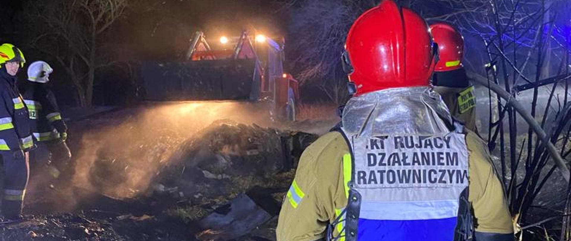Zdjęcie na pierwszym planie przedstawia strażaka odwróconego plecami do fotografa. Strażak w umundurowaniu specjalnym, w czerwonym hełmie na głowie oraz kamizelce, na której widnieje napis Kierujący Działaniem Ratowniczym. W tle ciągnik rolniczy z ładowaczem czołowym, który rozgarnia nadpaloną słomę i siano. Zdjęcie zrobione w porze nocnej.