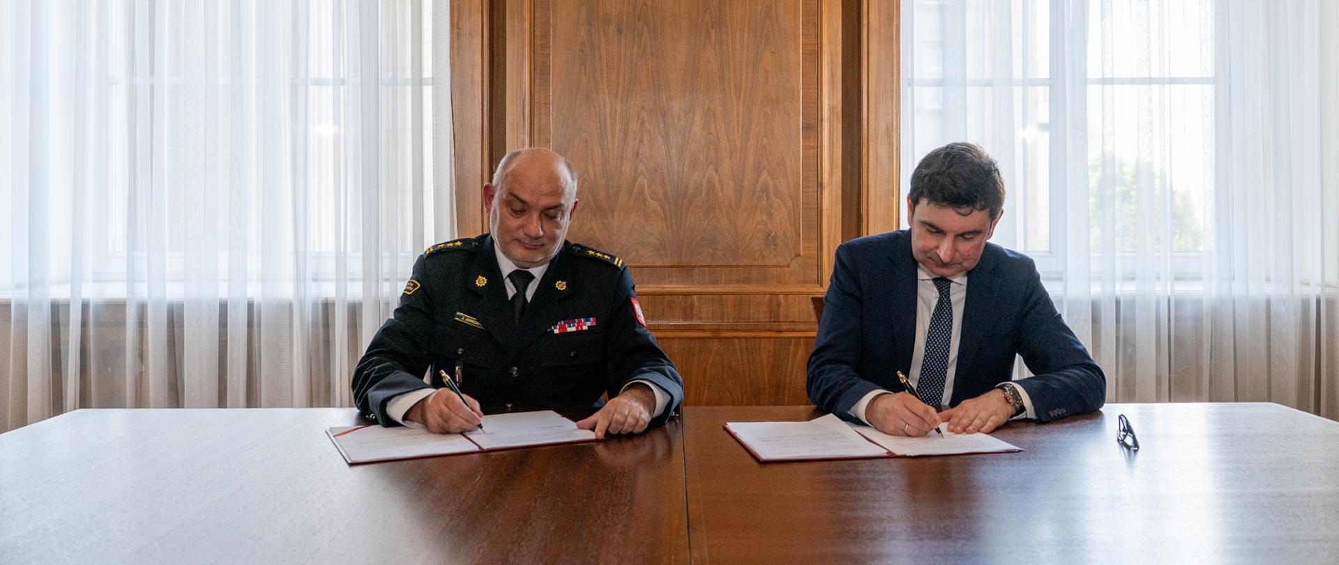 Wojewoda śląski Marek Wójcik oraz komendant Centralnej Szkoły Państwowej Straży Pożarnej w Częstochowie st. bryg. mgr inż. Jarosław Jankowski siedzą przy stole i podpisują umowę 