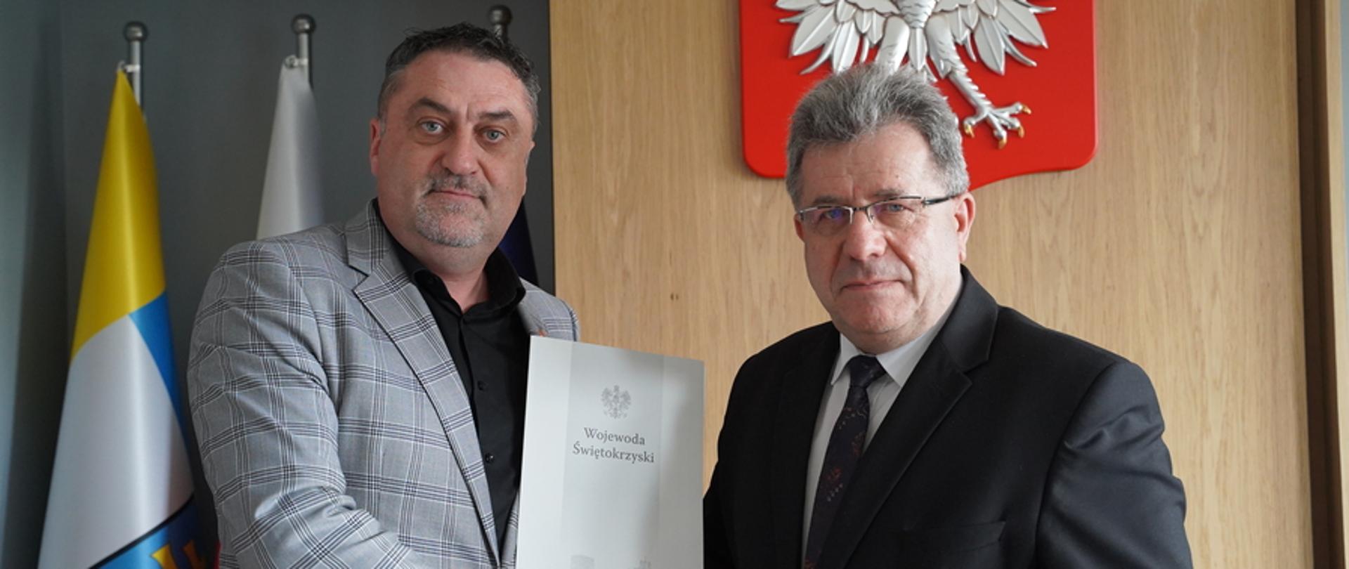 Wojewoda Józef Bryka stoi obok Piotra Łojka, podają sobie ręce, za nimi pod ścianą flagi Polski i Kielc, na ścianie wisi też godło państwowe.