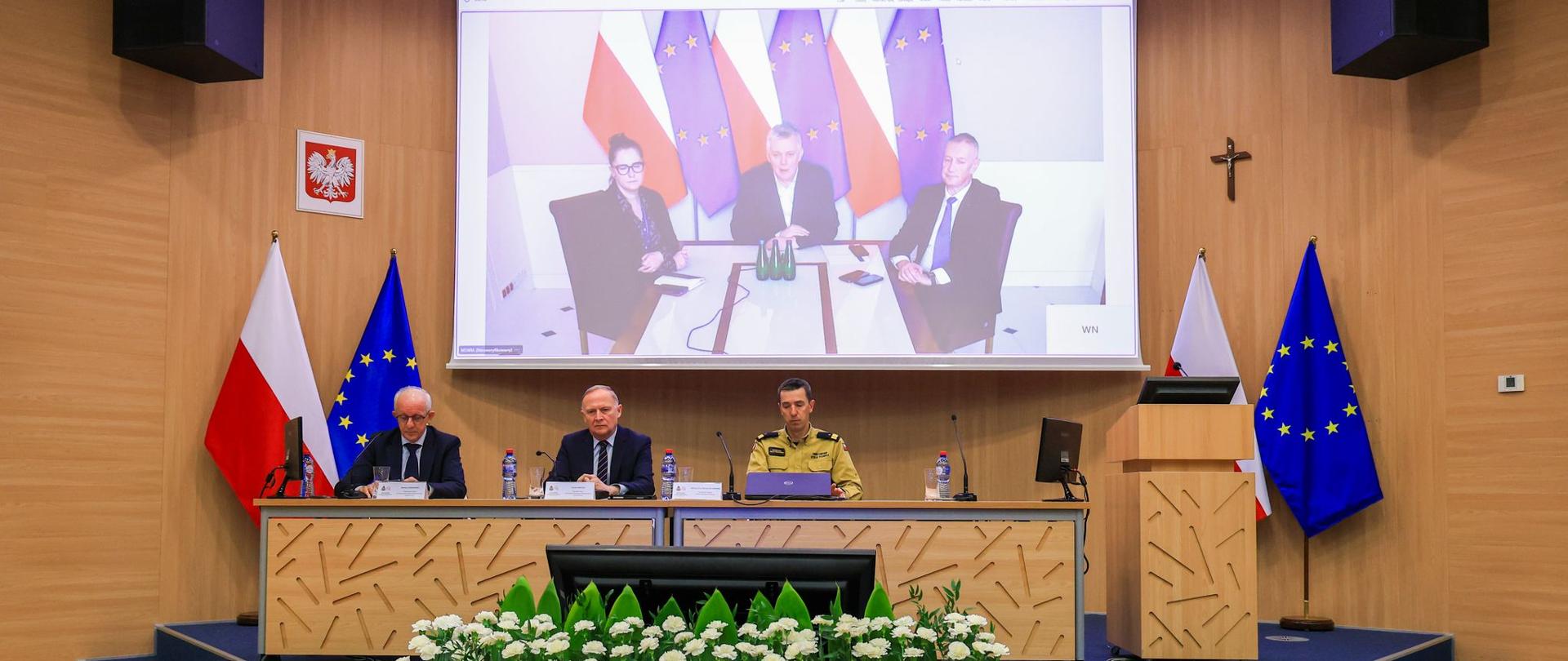 Trzech mężczyzn siedzi za stołem na podwyższeniu, dwóch wiceministrów w garniturach oraz jeden w mundurze nadbrygadiera Państwowej Straży Pożarnej. W tle na ekranie trzy osoby siedzą za stołem, dwóch mężczyzn i kobieta. Obok ekranu na stojakach stoją flagi Polski i Unii Europejskiej. Przed stołem na pierwszym planie kwiaty w kolorach białym i czerwonym ułożone na wzór flagi państwowej.