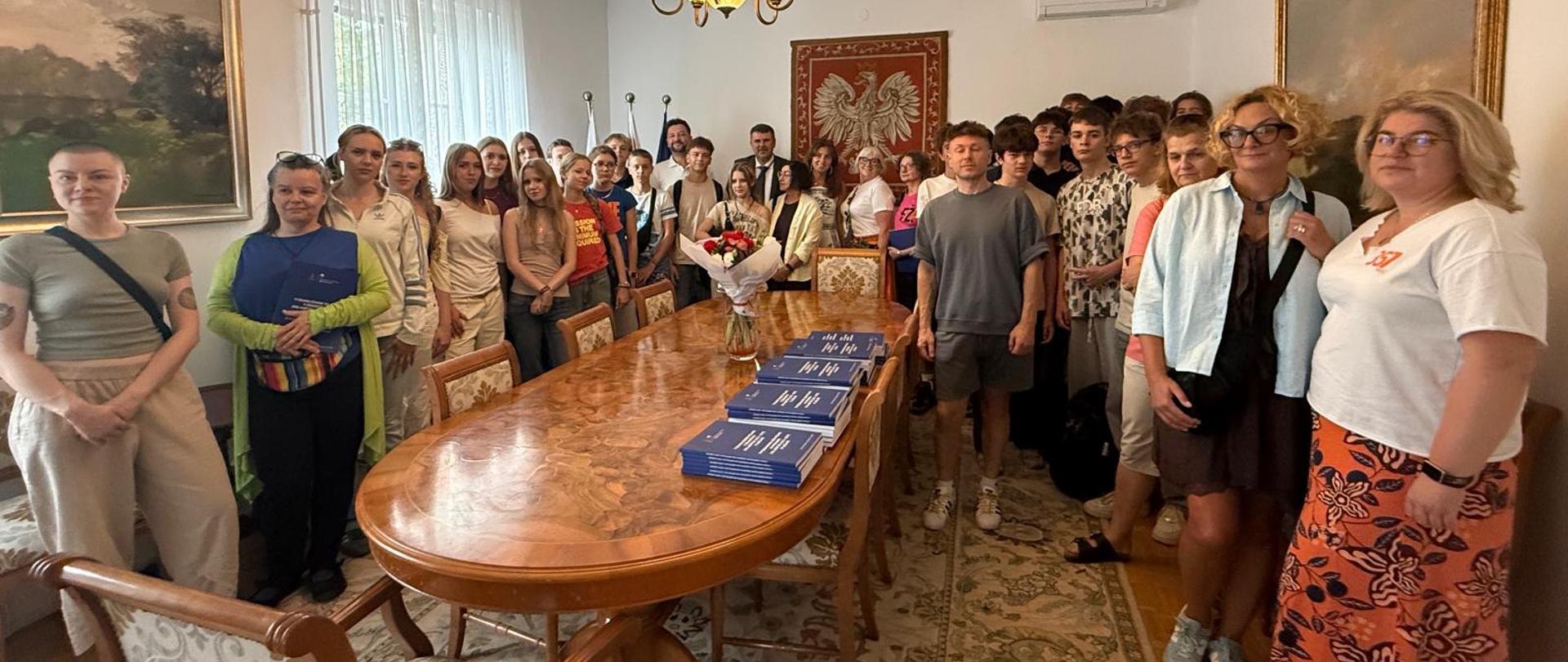 Duża grupa młodzieży i wykładowców pozuje do wspólnego zdjęcia w sali Ambasady RP w Lublanie. Uczestnicy stoją wokół długiego stołu, a na ścianie za nimi znajduje się godło Polski.