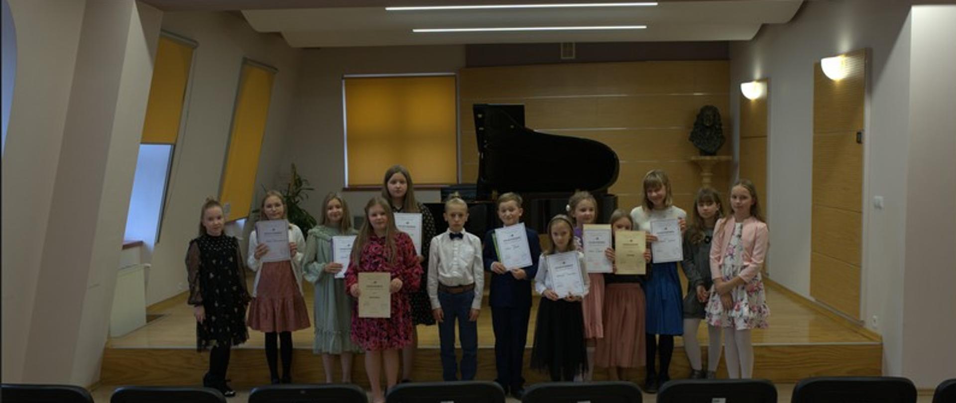 Zdjęcie przedstawia wszystkich uczestników konkursu stojących przed sceną z dyplomami. Przed dziećmi rząd krzeseł w tle fortepian.