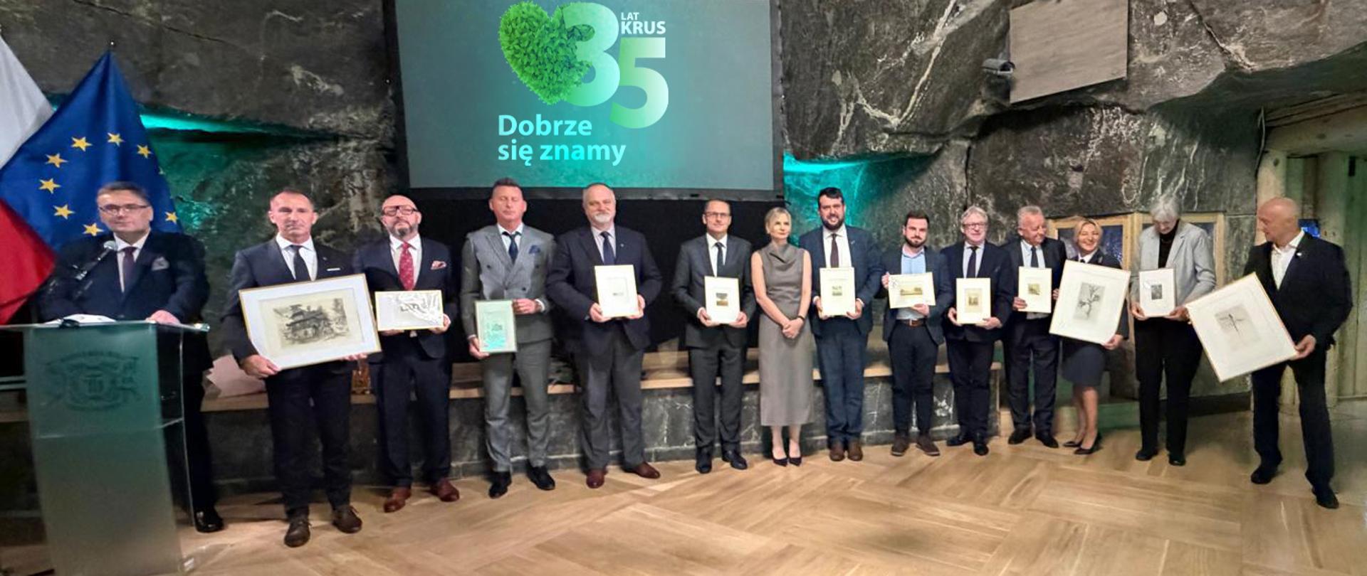 Prezes KRUS oraz zaproszeni goście podczas uroczystości z okazji 35-lecia KRUS w Krakowie. Uczestnicy pozują do wspólnego zdjęcia, trzymając pamiątkowe obrazy. W tle widoczne jubileuszowe hasło "35 lat KRUS. Dobrze się znamy"