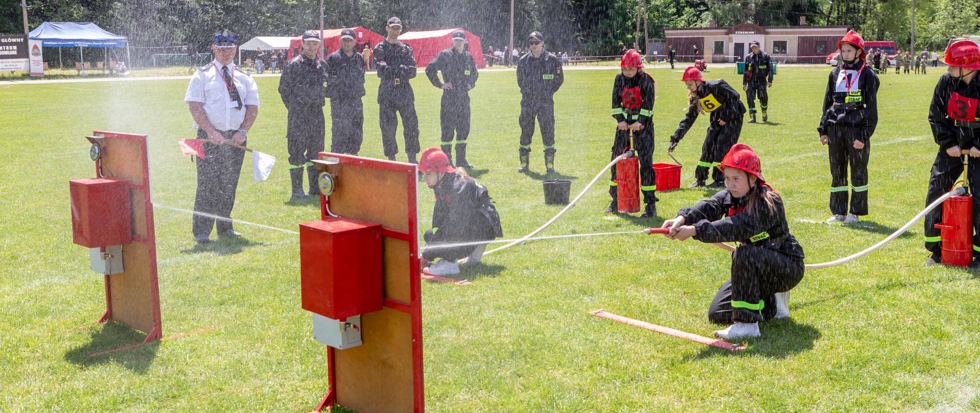 Zdjęcie grupę strażaków z Młodzieżowej Grupy Ochotniczej Straży Pożarnej w umundurowaniu ćwiczebnym. Po lewej stronie zdjęcia stoją nalewaki służące do gromadzenia wody podawanej przez prądownicę strażacką