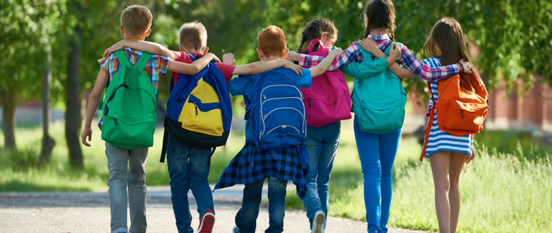 Na zdjęciu widać grupę sześciorga dzieci idących razem w plenerze, obejmujących się wzajemnie ramionami. Mają kolorowe plecaki i są zwrócone tyłem do aparatu. Scena przedstawia przyjaźń, współpracę i wspólne spędzanie czasu.
