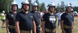 Zawody gminne w Śląskowie. Na placu ćwiczeń drużyna seniorów z Ochotniczej Straży Pożarnej w Ostojach. Występują w czarnych koszulkach z krótkim rękawem oraz z białych hełmach. W tle drzewa.