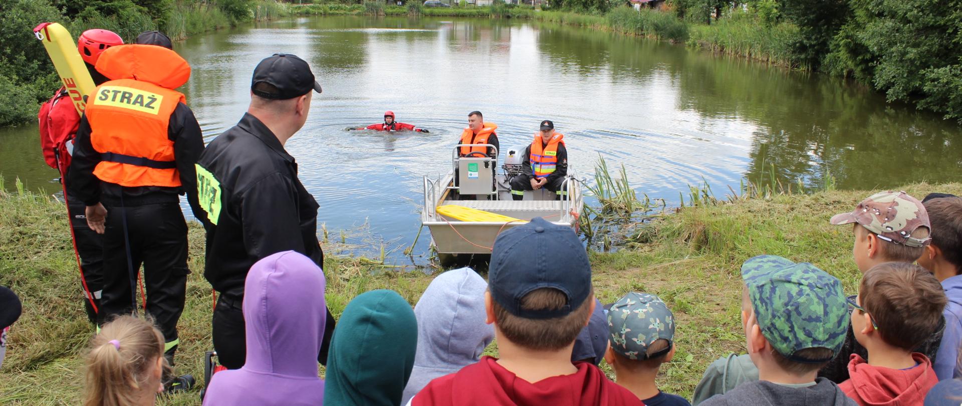 Dzieci oglądające strażacki pokaz ratownictwa wodnego. Przy brzegu strażacy w łodzi motorowej. W wodzie strażak pozorujący osobę tonącą. Obok, na brzegu strażacy uczestniczący w pokazach ubrani w kamizelkę ratunkową oraz skafander wypornościowy.