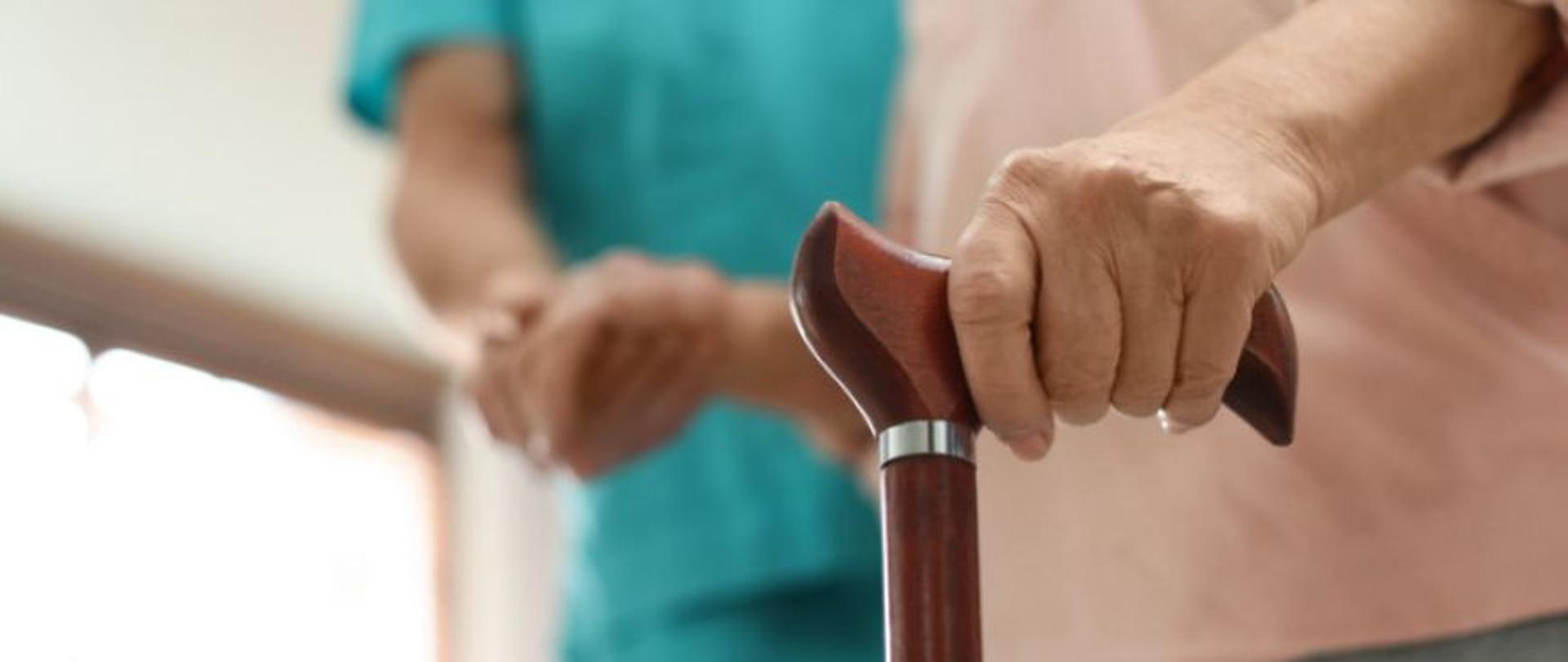 dwie osoby, jedna starsza trzyma w dłoni laskę, druga w pielęgniarskim stroju pomaga jej