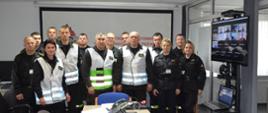 Zdjęcie przedstawia wszystkich uczestników ćwiczenia tworzących sztab KP Starogard Gdański. Strażacy w umundurowaniu i kamizelkach informacyjnych. w tle widać system wideokonferencyjny, na centralnej części zdjęcia stół z systemami łączności.