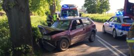 Zdjęcie przedstawia i bordowego Volkswagen Golf III uderzył w drzewo, który uderzył w drzewo. Po lewej stronie pojazdu stoi radiowóz policyjny zaś za rozbitym autem karetka pogotowia.
