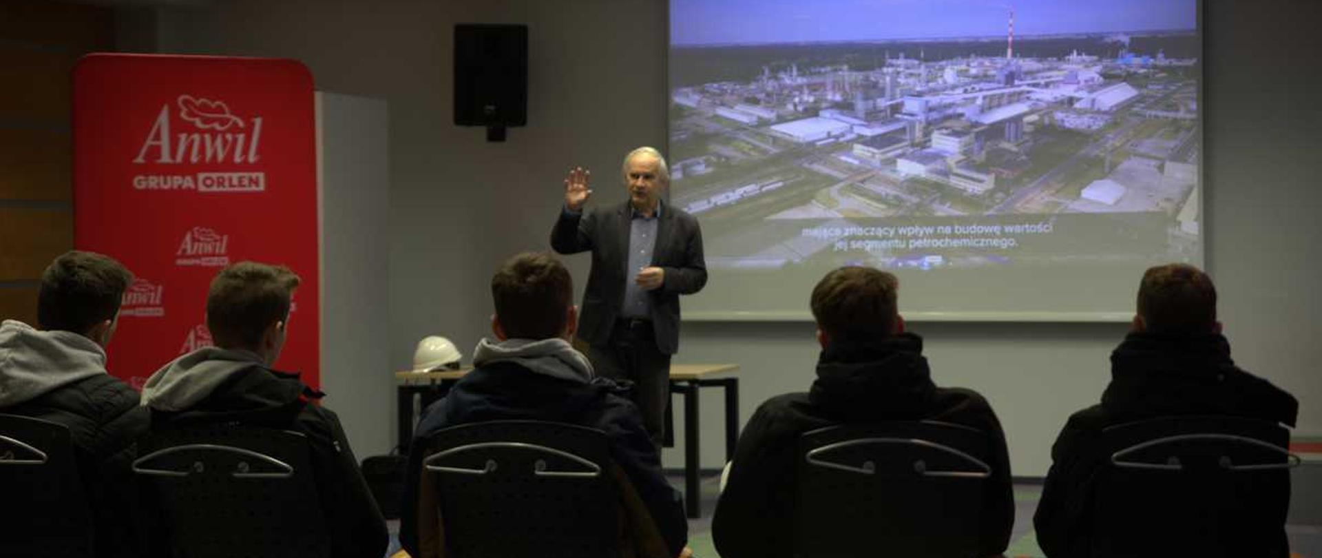 Zdjęcie przedstawia grupę osób w sali konferencyjnej podczas wykładu. Na tle wykładowcy widać ekran wyswietlający zakłady chemiczne we Wlocławku.