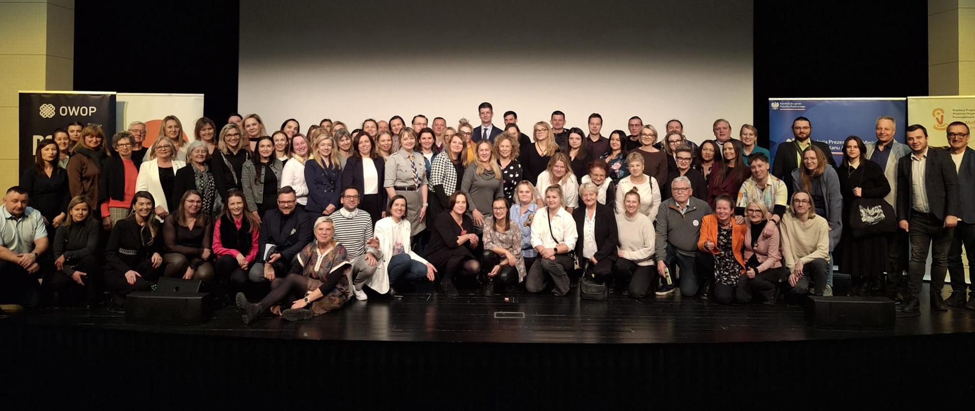Zdjęcie grupowe. Szeroki kadr, widok obejmuje scenę. Uczestnicy wydarzenia pozują do wspólnego zdjęcia. 