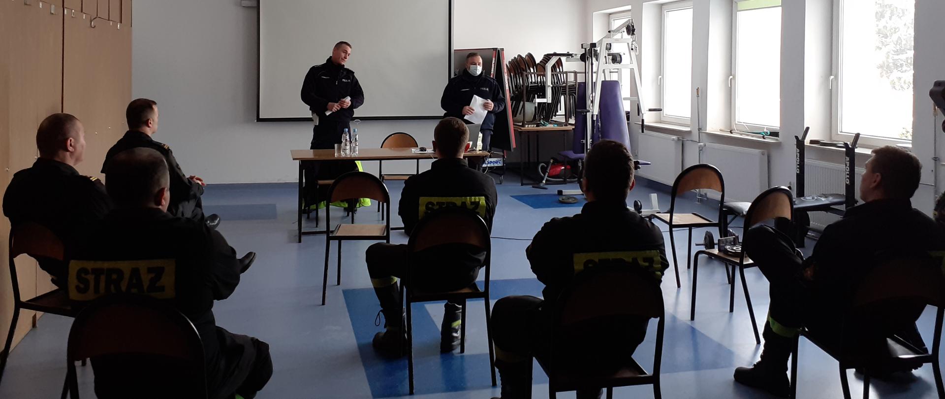 Na pierwszym planie widać siedzących tyłem na krzesłach strażaków. W dalszej części widać dwóch stojących policjantów, przed którymi ustawiony jest stolik, a na nim komputer. Wszyscy znajdują się w pomieszczeniu budynku.