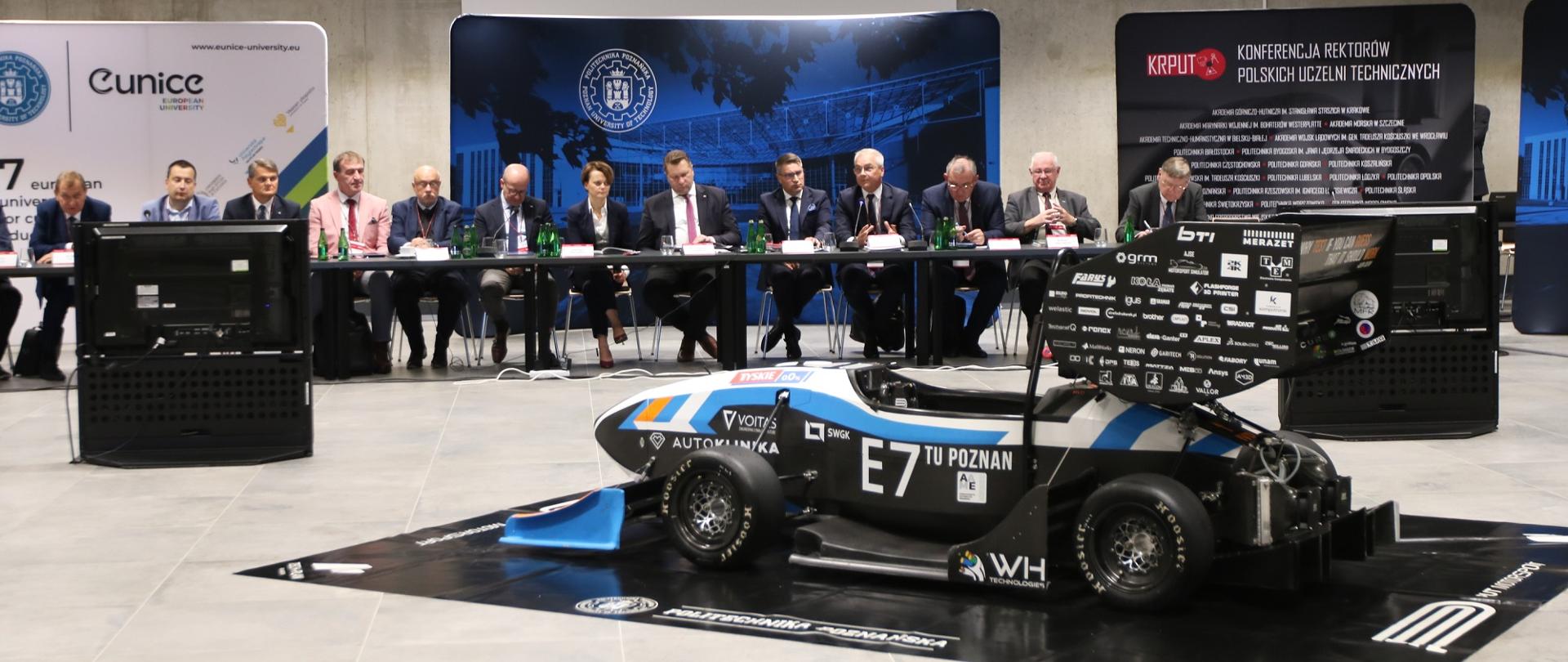 Przy szarym stole siedzi minister Czarnek i kilka innych osób, przed stołem na podłodze stoi mały, niski, czarny z niebiesko-białym paskiem samochód z napisem E7 TU Poznań.