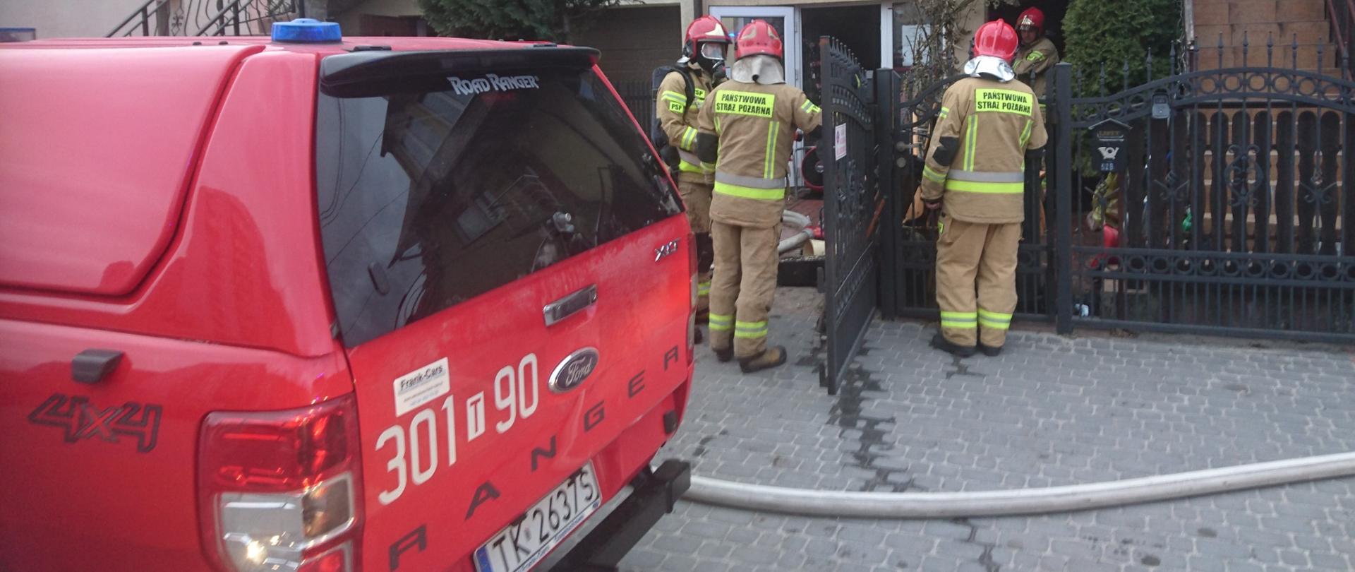 Fotografia przedstawia ratowników w czasie trwania akcji gaśniczej, na chodniku leży nawodniona linia gaśnicza, na pierwszym planie fragment samochodu pożarniczego. Tłem jest budynek, w którym wybuchł pożar.