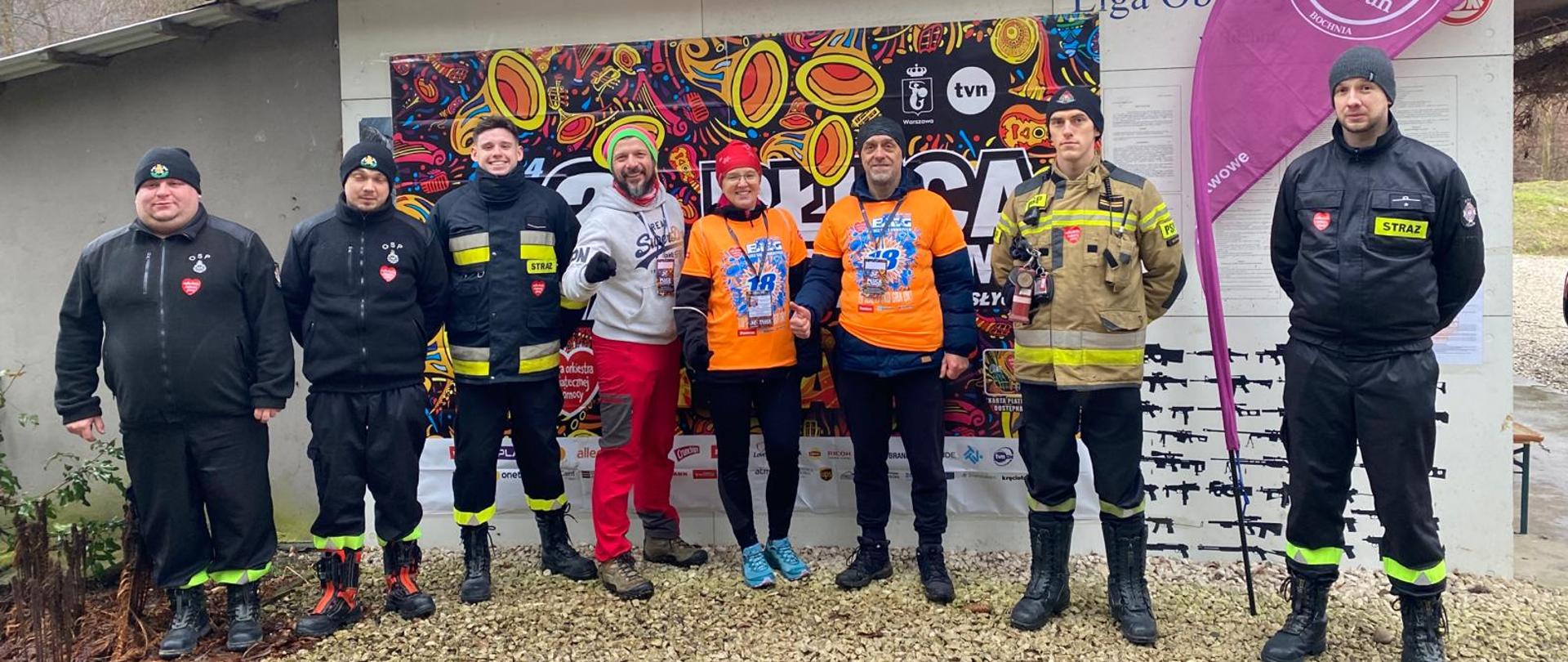 Kilku mężczyzn ubranych w czarne ubrania koszarowe strażackie z odblaskowymi paskami, czarnym butami oraz trzema mężczyznami ubranymi w strój sportowy stoją polu przed budynkiem. W tle budynek i napis wielkiej orkiestry świątecznej pomoc