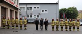 Zdjęcie przedstawia Komendanta Powiatowego Państwowej Straży Pożarnej w Zambrowie starszego brygadiera Krzysztofa Konieckiego w mundurze wyjściowym, Zastępcę Komendanta Powiatowego Państwowej Straży Pożarnej w Zambrowie brygadiera Adama Wysmułka w mundurze wyjściowym oraz nowo mianowanego Dowódcę Jednostki Ratowniczo-Gaśniczej w Zambrowie starszego kapitana Pawła Gwardiaka w mundurze wyjściowym pozujących do zdjęcia w szeregu ze strażakami z Jednostki Ratowniczo-Gaśniczej w Zambrowie ubranymi w ubranie bojowe z hełmami. Zdjęcie wykonane na placu Komendy Powiatowej Państwowej Straży Pożarnej w Zambrowie, na tle części budynku.