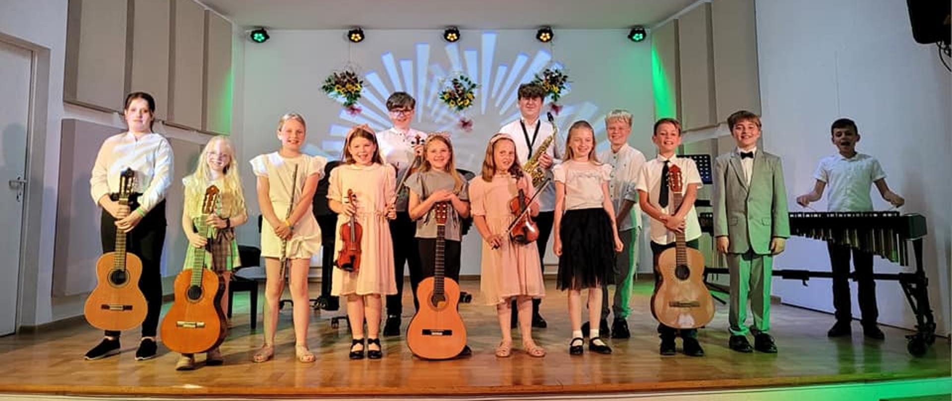 Na zdjęciu widać uczniów klas pierwszych, którzy stoją na scenie trzymając różne instrumenty