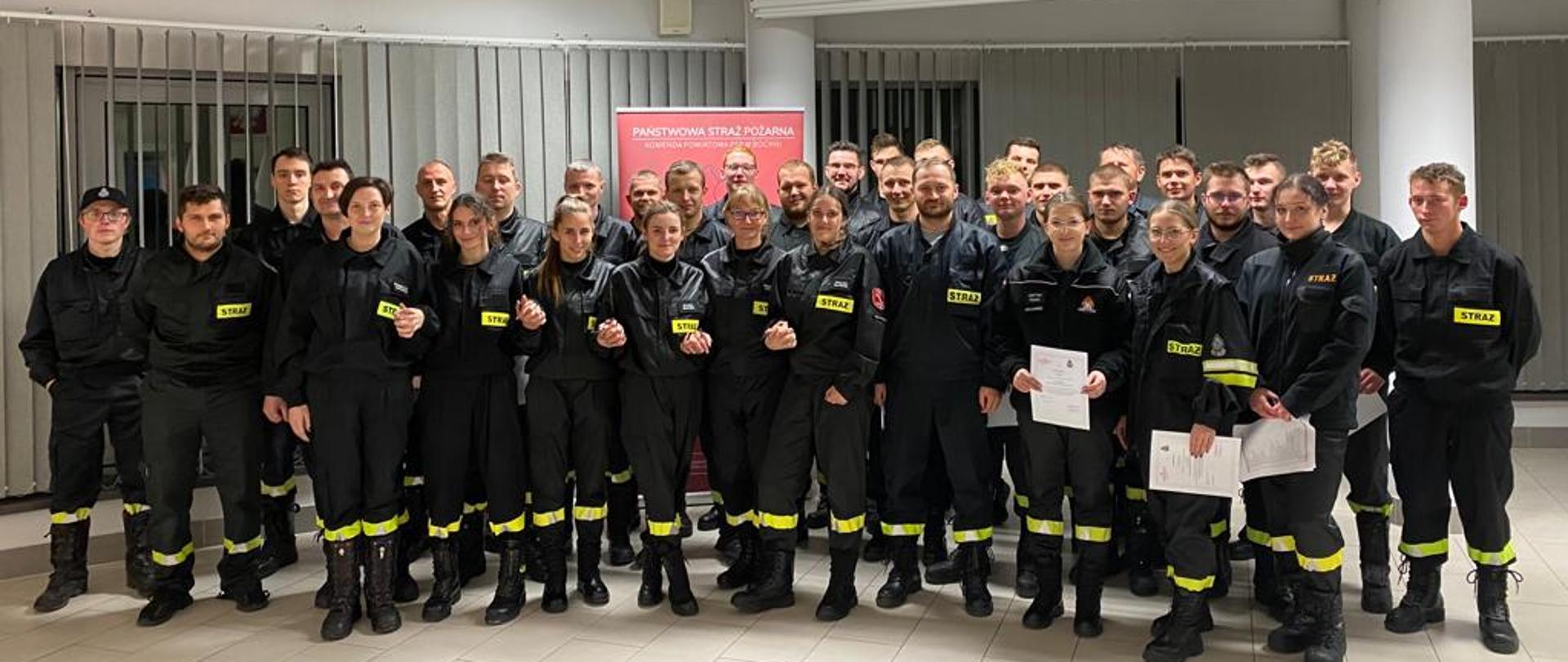 Kilkudziesięciu mężczyzn ubranych w czarne ubrania koszarowe strażackie z odblaskowymi paskami, czarnym butami stoją na Sali konferencyjnej. W tle ściany w kolorze białym, żaluzje koloru szarego