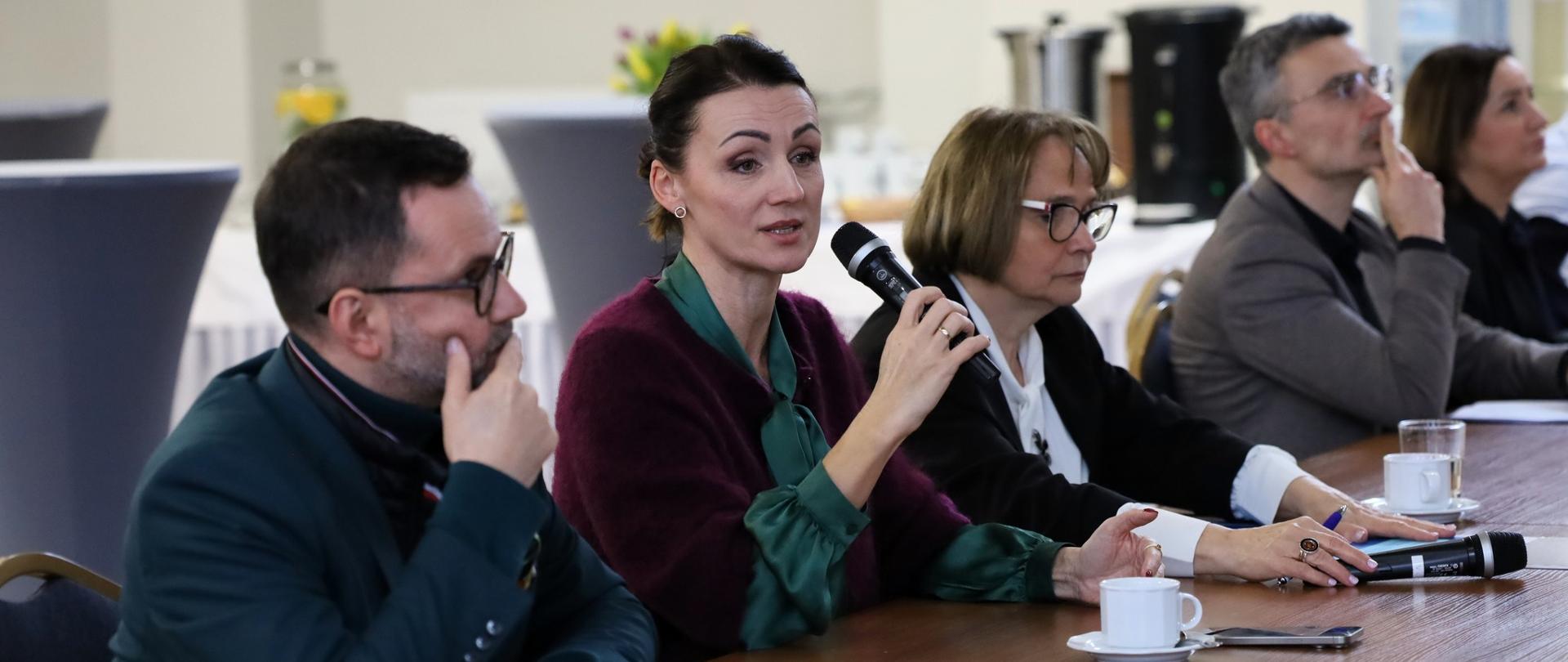 kilka osób siedzi przy stole, jedna (wojewoda) trzyma w ręce mikrofon i przemawia