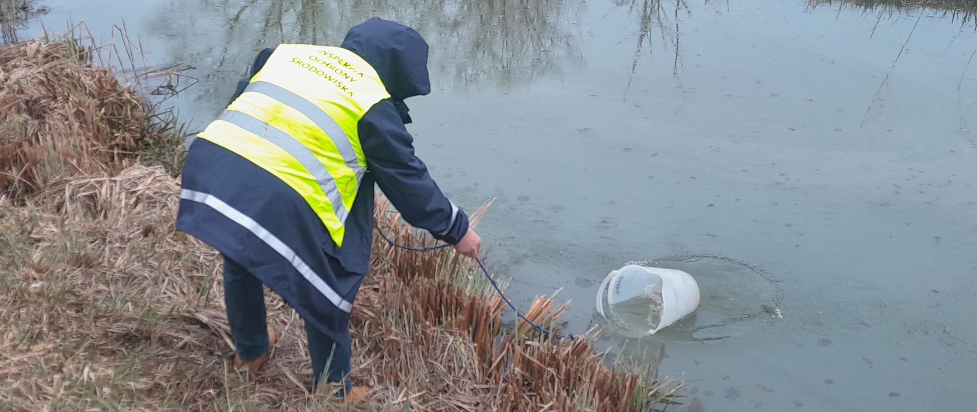 Pobór próbek przez CLB GIOŚ. Próbkobiorca w żółtej kamizelce pobiera wodę z rzeki. W tle ogródki działkowe.