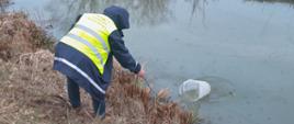 Pobór próbek przez CLB GIOŚ. Próbkobiorca w żółtej kamizelce pobiera wodę z rzeki. W tle ogródki działkowe.