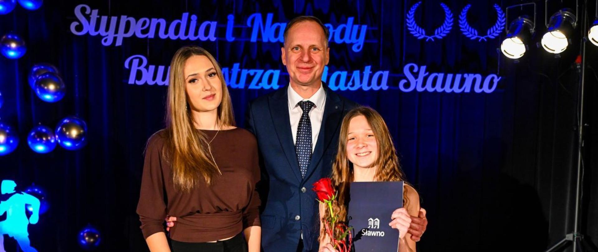 Zdjęcie kolorowe. Dwie kobiety i mężczyzna pozujący do wspólnej fotografii na scenie. Mężczyzna pośrodku ubrany w ciemny garnitur, białą koszulę i krawat w kropki. Kobieta po lewej stronie w brązowej bluzce z długim rękawem i czarnych spodniach. Kobieta po prawej stronie w czarnej sukience, trzymająca w dłoniach czerwoną różę oraz ciemnoniebieski folder z napisem "Sławno". W tle ciemnoniebieska kurtyna z białym napisem "Stypendia i Nagrody Burmistrza Miasta Sławno" oraz dekoracyjne niebieskie kule i podświetlona figura sportowca.