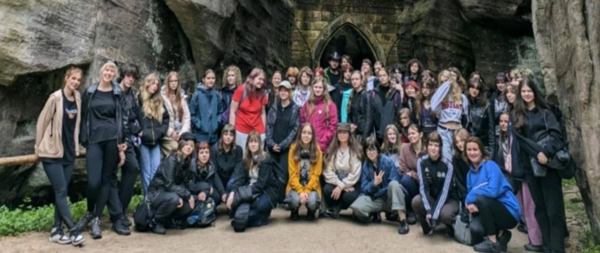 Zdjęcie wykonano w Pradze, a atmosfera jest luźna i koleżeńska. Wszyscy pozują z uśmiechem, nieformalnie, w swobodnych pozach.
Grupa około 50 młodych osób pozuje do wspólnego zdjęcia grupowego na tle monumentalnych formacji skalnych. Uczestnicy stoją i kucają na ścieżce między wysokimi, wąsko rozstawionymi ścianami skalnymi, które tworzą naturalny wąwóz. Skały mają pionowe warstwowania i są w odcieniach szarości, brązu i zieleni — miejscami porośnięte mchem lub roślinnością. U podstawy jednej ze skał znajduje się kamienny portal o łukowatym kształcie, przypominający wejście do groty lub tunelu – stylizowany na gotycki, z mchem i śladami erozji.
Osoby na zdjęciu są ubrane w odzież turystyczną lub sportową – kurtki, bluzy, spodnie trekkingowe. Część siedzi na ziemi lub przysiadła, reszta stoi. W grupie widać dużą różnorodność stylów i kolorów ubrań – od stonowanych czerni i szarości po żywe kolory, jak żółty, czerwony czy niebieski. Niektórzy noszą czapki lub kaptury. Atmosfera jest swobodna i pogodna – wiele osób uśmiecha się do aparatu.
Zdjęcie wykonano w tzw. „Skalnym Mieście” – obszarze przyrodniczym z unikalnymi formacjami skalnymi, możliwe że w Adršpašsko-Teplickich Skałach w Czechach.