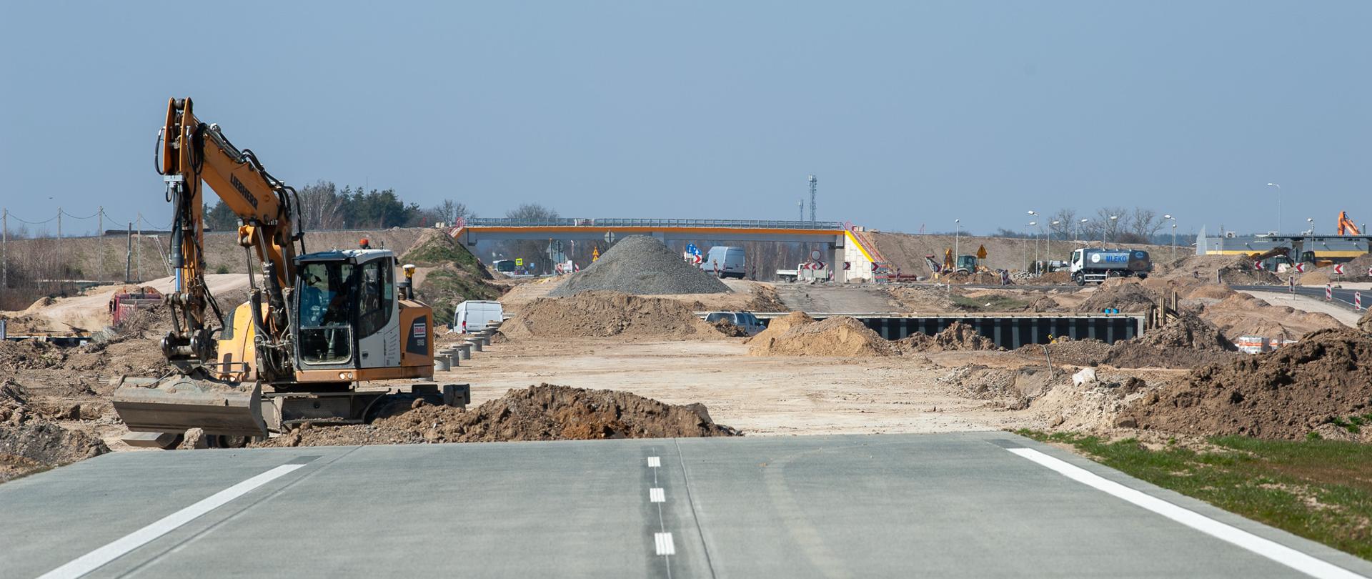 Na zdjęciu widoczna jest nieukończona jezdnia z wyraźnie zaznaczonymi pasami ruchu. W tle znajduje się koparka pracująca nad usuwaniem ziemi oraz inne maszyny budowlane i materiały, takie jak stosy piasku i żwiru. 