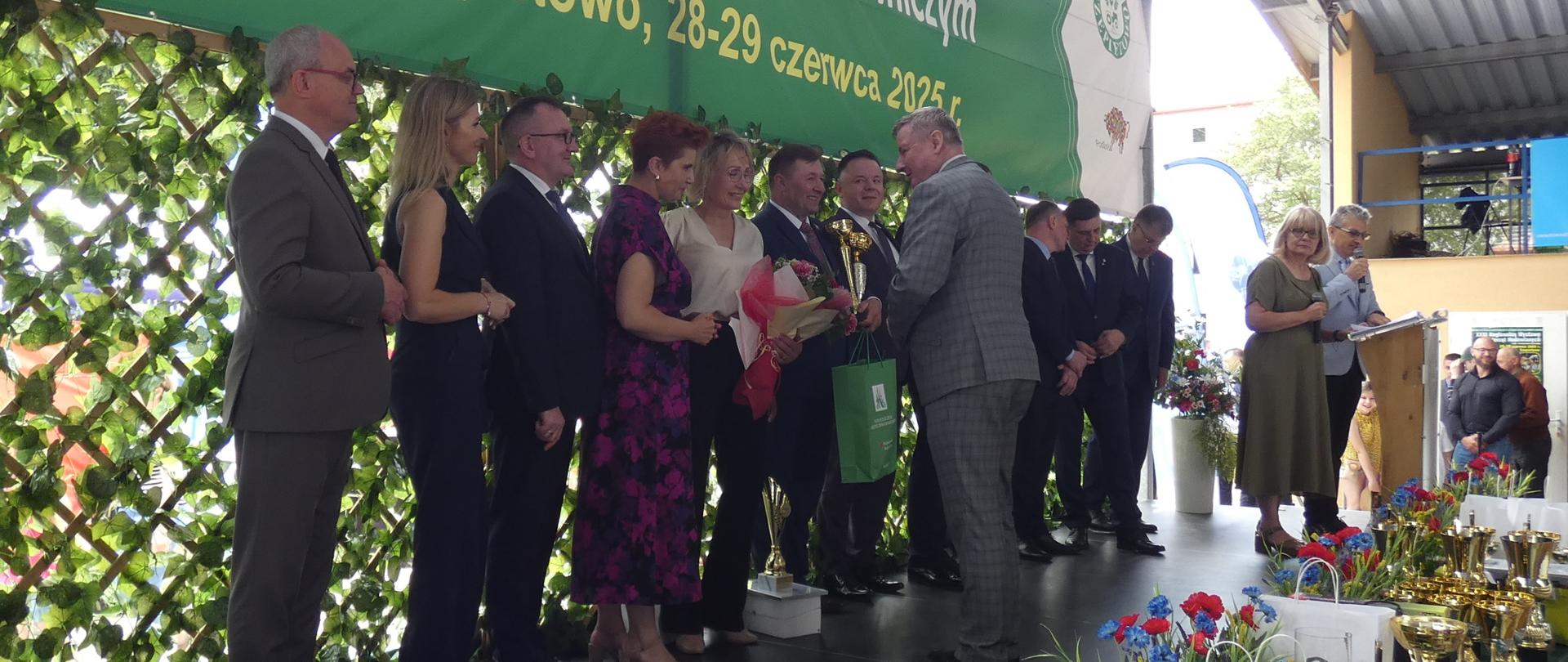 Na scenie pod zadaszeniem trwa uroczystość wręczenia nagród podczas Regionalnej Wystawy Zwierząt Hodowlanych i Dni z Doradztwem Rolniczym w Szepietowie. Zastępca Dyrektora Oddziału Regionalnego KRUS w Białymstoku Jarosław Borowski, w eleganckim, szarym garniturze, wręcza nagrodę Dorocie i Bogusławowi Ciupińskim, Mistrzom Podlaskiej Agroligi 2024 w kategorii „Rolnik”. Obok niego stoi grupa zaproszonych gości i nagrodzonych osób, w tym kobieta trzymająca bukiet kwiatów. W tle widoczny jest zielony baner z nazwą wydarzenia, a na pierwszym planie ustawiono liczne puchary, torby z nagrodami i bukiety kwiatów.