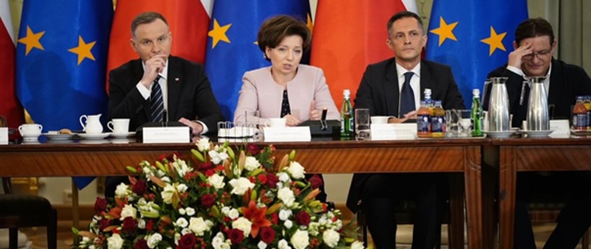 Zdjęcie zrobione w sali Kancelarii Prezydenta RP. Za stołem prezydialnym siedzą od lewej strony: Andrzej Duda - Prezydent RP, Marlena Maląg - Minister Rodziny i Polityki Społecznej, Łukasz Bernatowicz - Przewodniczący Rady Dialogu Społecznego i Jacek Męcina - Konfederacja Lewiatan. Przed stołem, po środku na podłodze stoi bukiet kwiatów w kolorze biało-czerwonym. W tle siedzących widnieje pięć flag: UE, Polski i tak na przemian. Na stole znajdują się mikrofony, kawa herbata.