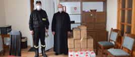 Na zdjęciu widać strażaka w ubraniu koszarowym, obok niego stoi ksiądz. Mężczyźni mają usta i nos zasłonięte maseczkami ochronnymi. Na podłodze obok mężczyzn są ułożone papierowe kartony i baniaki z płynem do dezynfekcji.