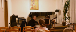 Zdjęcie przedstawia dwa rzędy krzeseł oraz widownię, przed którą wykładowca objaśnia pewne zagadnienia uczniowi grającemu na gitarze. W tle na scenie znajduje się czarny fortepian oraz portret patronki szkoły muzycznej w Oławie.