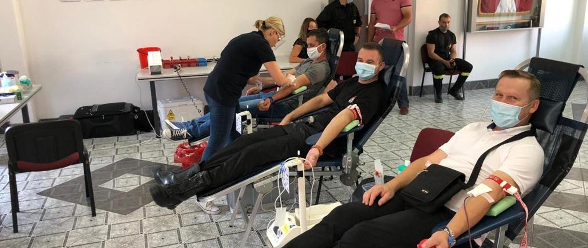 Pomieszczenie, wewnątrz którego stoi trzy czarne fotele na metalowych stelażach. Na fotelach, w pozycji siedząco-leżącej znajdują się mężczyźni. Mężczyzna na pierwszym planie w lewej ręce posiada wkłucie, którym pobierana jest krew, dwaj pozostali mężczyźni są przygotowywani do zabiegu - przy jednym z nich stoi kobieta. Pod ścianą, na której widoczny jest sztandar oraz dyplomy sportowe, stoi grupa osób.
. Za fotelami, przy ścianie, pod oknem widoczny stolik, przy którym siedzi kobieta i mężczyzna, obok stolika stoi inna kobieta, która trzyma mężczyznę za ramię. Pod ścianą na krześle siedzi kobieta. Na ścinie widoczne sportowe trofea oraz puchary. 