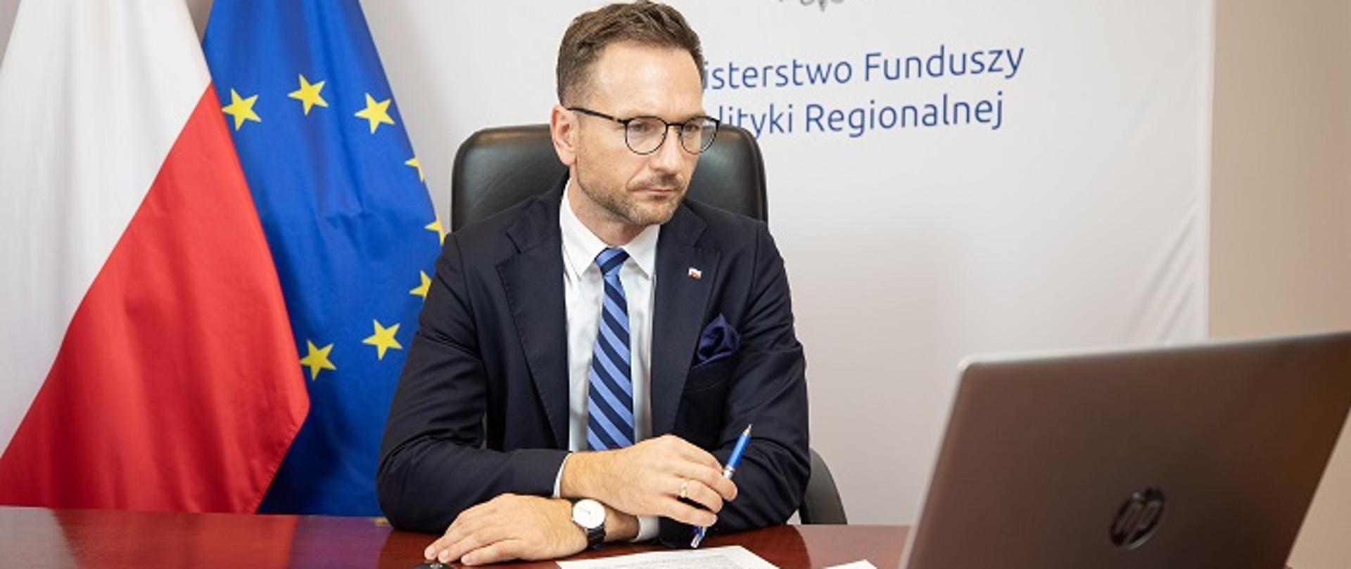 Wiceminister Waldemar Buda siedzi przy biurku, przed laptopem. Z tyłu widoczne flagi Polski i UE oraz logo MFiPR.