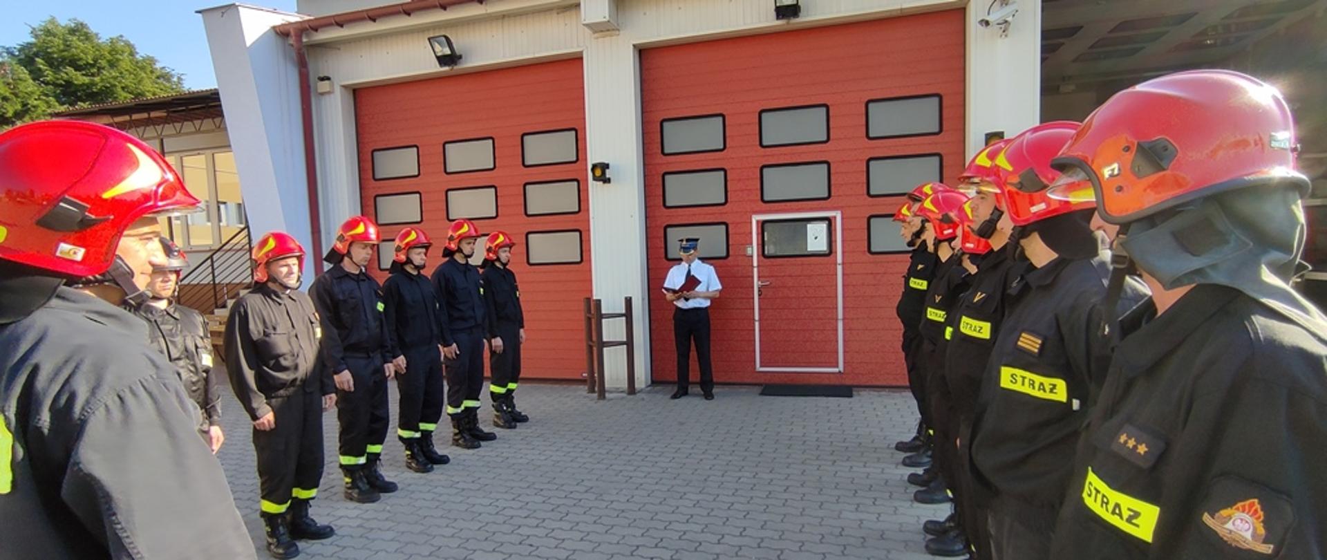 Plac wewnętrzny komendy, strażacy ustawieni naprzeciw siebie w dwóch szeregach z zachowaniem odstępów, trwa codzienna zmiana służby w jednostce ratowniczo-gaśniczej, komendant powiatowy PSP odczytuje treść dyplomu