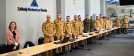 Za stołami ustawionym wzdłuż dużego pomieszczenia stoją uczestnicy ćwiczeń, strażacy w mundurach piaskowych, żołnierze w ubraniach typu moro. Na ścianie logo Zakładów Mechanicznych, oraz fototapeta z rodzajami broni palnej produkowanej na ZM