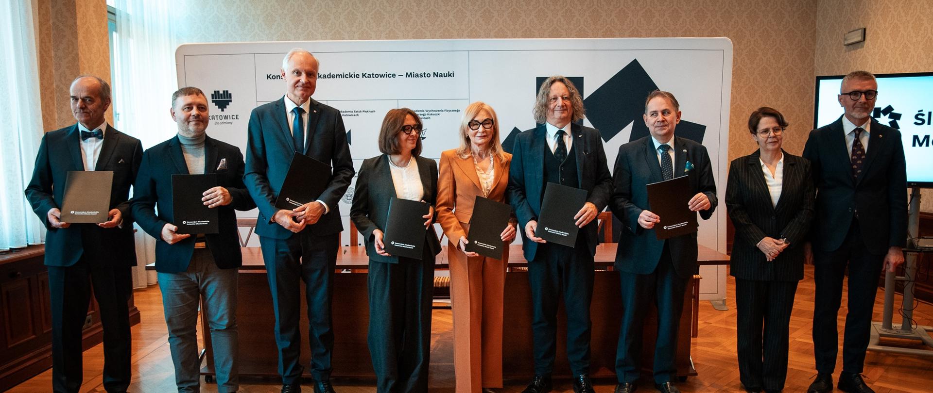 W sali z drewnianą podłogą w szeregu stoi dziewięć osób, trzymają czarne teczki, za nimi drewniane biurko, z tyłu pod ścianą biała ścianka z napisem Konsorcjum Akademickie Katowice - Miasto Nauki, z lewej zasłonięte firankami okna.