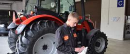 Uczeń w stroju roboczym stoi przedciągnikiem, przegląda książkę serwisową