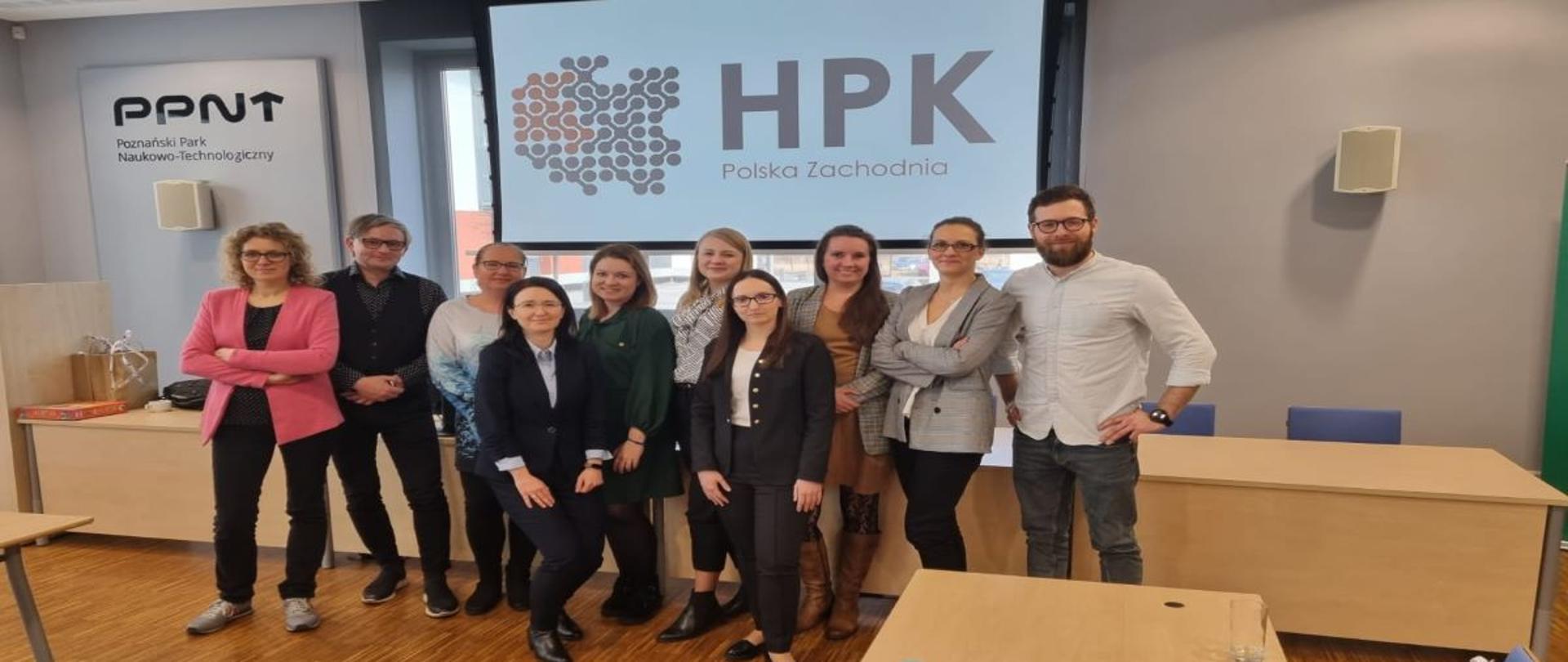 Grupa dziesięciu osób stoi razem obok siebie. W grupie jest dwóch mężczyzn i osiem kobiet. Za grupą u góry na ścianie jest wyświetlone logo Horyzontalnego Punktu Kontaktowego Polska Zachodnia. Po lewej stronie na ścianie umieszczone jest logo Poznańskiego Parku Naukowo-Technologicznego. Za osobami na ziemi jest długie drewniane biurko.