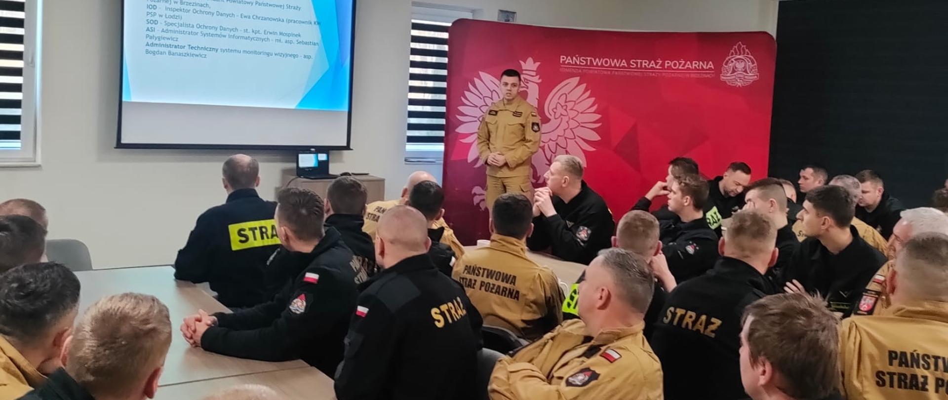 Odprawa służbowa , w której uczestniczył Komendant Powiatowy Państwowej Straży Pożarnej w Brzezinach starszy brygadier Mariusz Kaczmarski, Zastępca Komendanta Powiatowego Państwowej Straży Pożarnej w Brzezinach młodszy brygadier Jakub Brzózka oraz funkcjonariusze i pracownicy cywilni komendy. Załoga siedzi przy stołach, dowódca Jednostki Ratowniczo-Gaśniczej stoi przy ekranie i referuje zagadnienia z zakresu ochrony danych osobowych, wyświetla slajdy na ekranie, za nim widoczna ścianka reklamowa komendy. 