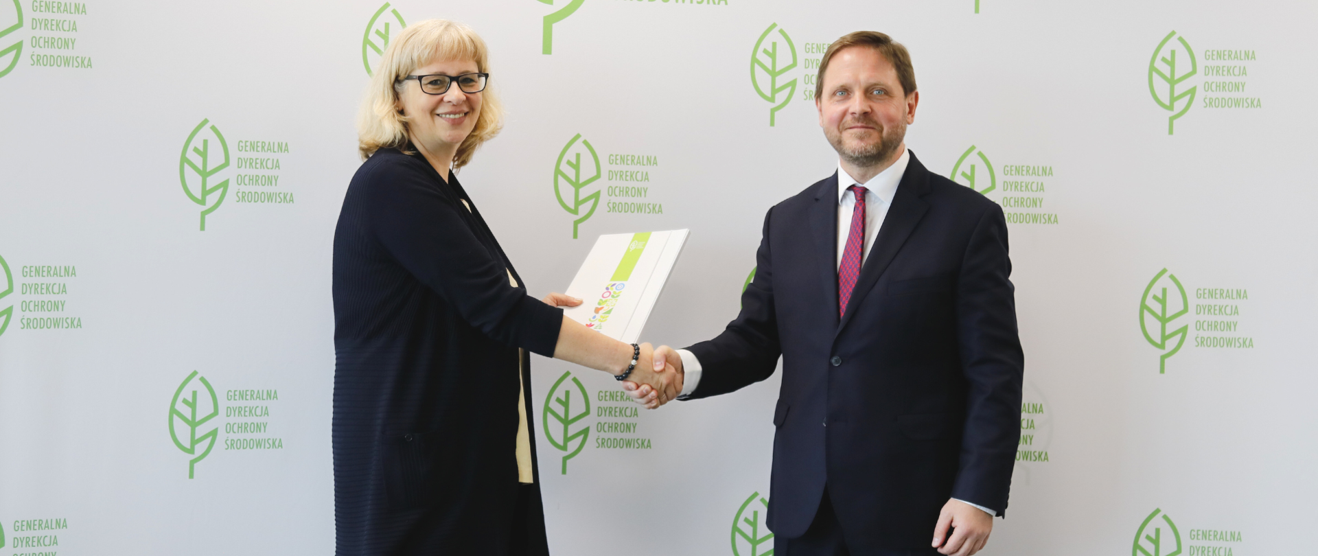 Na tle białej ściany z logotypami Generalnej Dyrekcji Ochrony Środowiska stoją uśmiechnięta kobieta i mężczyzna, ściskają sobie dłonie. Kobieta w drugiej dłoni trzyma teczką.