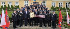 Druhów OSP, strażaków oraz wiceministra którzy stoją na schodach jeden za drugim i pozują do zdjęcia. Ubrani są w umundurowania wyjściowe. Trzy osoby w rękach trzymają obraz i szable pamiątkowe.