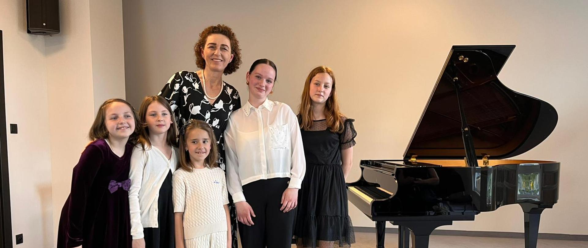 Na zdjęciu widzimy sześć osób - nauczycielka z pięcioma uczennicami stojących obok czarnego, lśniącego fortepianu koncertowego, który znajduje się po prawej stronie kadru. Tłem jest jasna ściana.
Po lewej stronie, lekko z tyłu, stoi kobieta o kręconych, ciemnych włosach, ubrana w elegancką, czarno-białą sukienkę we wzory. Uśmiecha się. Wygląda na nauczycielkę lub opiekunkę młodych dziewcząt.
Przed nią, na pierwszym planie, stoi pięć dziewcząt w różnym wieku, ubrane odświętnie.
Od lewej do prawej:
Pierwsza dziewczynka, najniższa z lewej strony, ma na sobie ciemnofioletową, aksamitną sukienkę i białe rajstopy. Jej ciemne włosy są rozpuszczone.
Obok niej stoi kolejna dziewczynka w czarnej sukience z białymi mankietami i kołnierzykiem, z ciemnymi, prostymi włosami.
Trzecia dziewczynka, również z ciemnymi włosami, ubrana jest w jasny, kremowy sweterek z warkoczami i jasne spodnie.
Czwarta dziewczynka, nieco starsza, stoi wyżej, ubrana w białą, elegancką koszulę z długimi rękawami i czarne spodnie. Ma jasne, proste włosy.
Ostatnia dziewczynka, po prawej stronie, obok fortepianu, ma na sobie czarną, koronkową sukienkę. Jej rude włosy są rozpuszczone.
Wszystkie dziewczynki są uśmiechnięte i patrzą w stronę obiektywu. Fortepian po prawej stronie kadru jest otwarty, ukazując struny i wewnętrzne elementy. Całość sprawia wrażenie uroczystego i radosnego wydarzenia, prawdopodobnie po występie muzycznym.