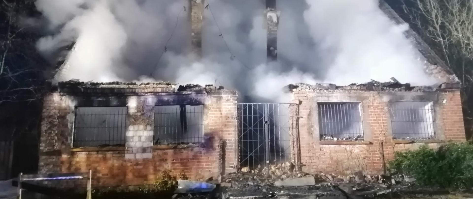 Fotografia przedstawia ujęcie frontu budynku leśniczówki. Ściany z czerwonej cegły, brak więźby dachowej, w otworach okiennych i drzwiowych kraty, z wnętrza budynku unosi się gęsty biały dym, wszędzie widać osmalenia po pożarze. Jest noc, na ziemi widoczne odcinki węży pożarniczych.