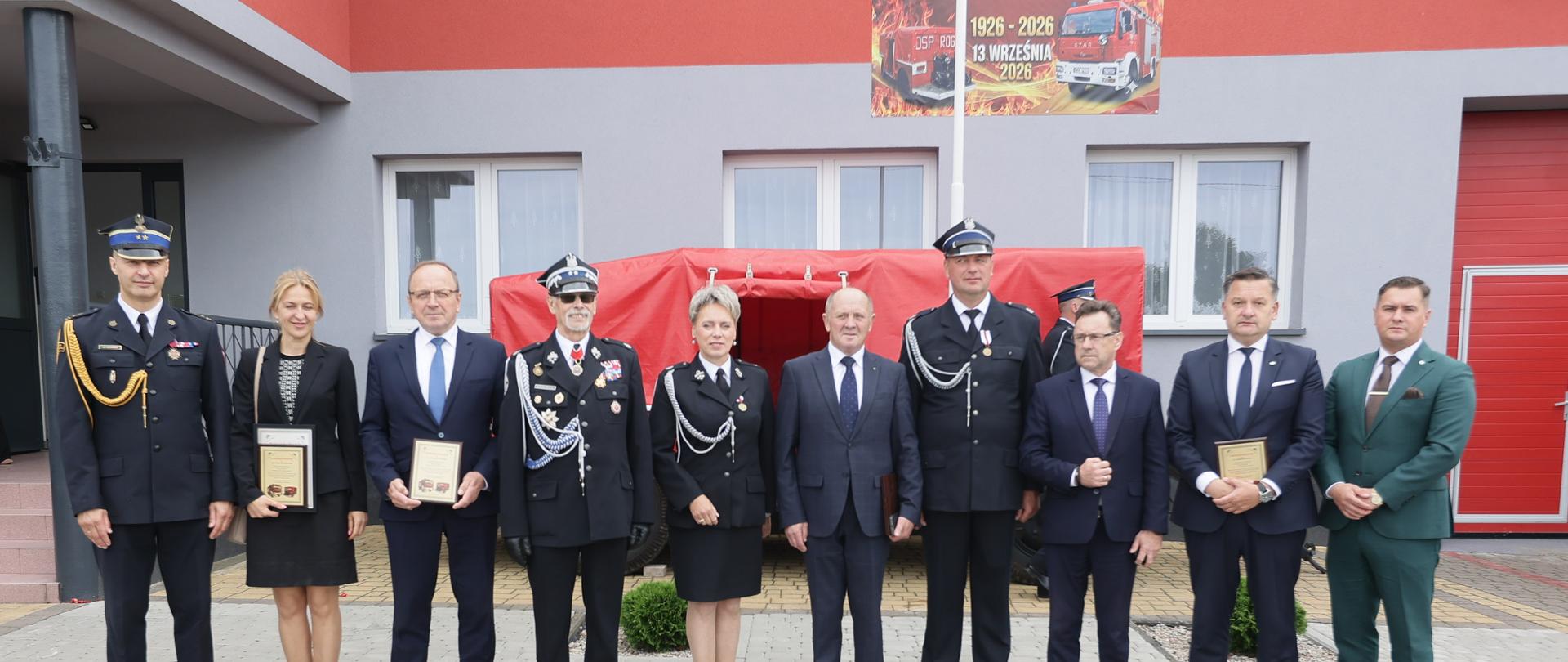 Pamiątkowe zdjęcie oficjalne na tle budynku remizy z banerem „100-lecie OSP Rogów 1926-2026”, na którym pozuje grupa zaproszonych gości, samorządowców oraz przedstawicieli służb trzymających pamiątkowe tabliczki.