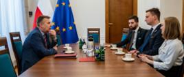 Zdjęcie z boku, minister Wieczorek siedzi przy stole i rozmawia z siedzącymi naprzeciwko dwoma mężczyznami w garniturach i kobietą w białej bluzce.